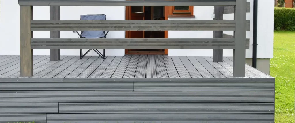 Part of white residential house with brown doors, windows and dark gray or anthracite wpc composite material terrace deck with wooden railings and skirting in backyard outdoors