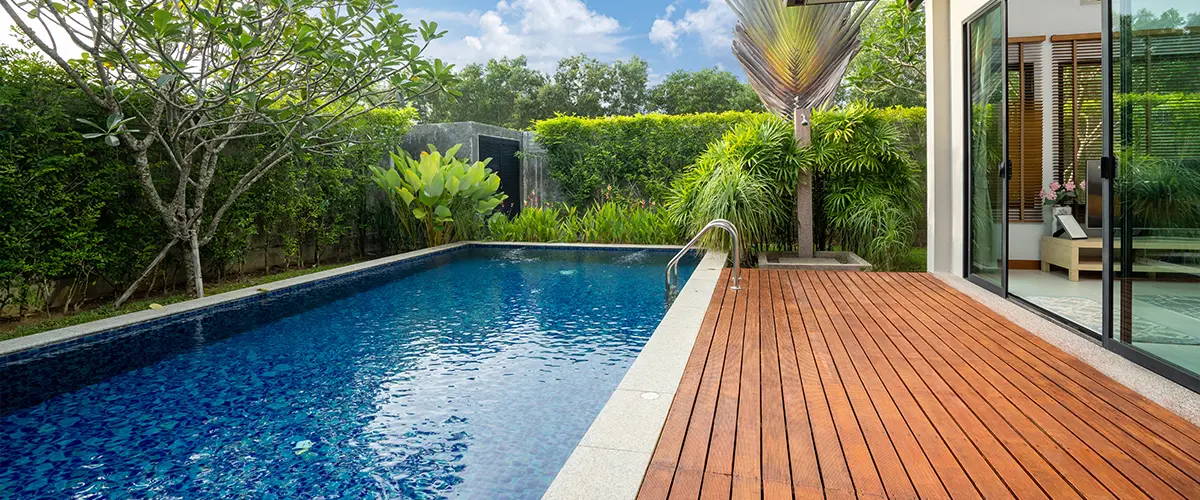 Modern luxury house backyard with blue swimming pool, wooden deck, and lush greenery on a sunny day.