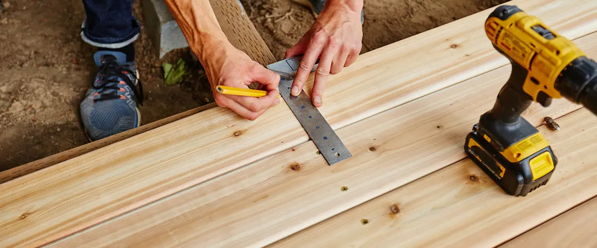 Carpenter measuring wooden deck boards with a square and pencil, alongside a cordless drill, demonstrating precision in deck installation in Knoxville