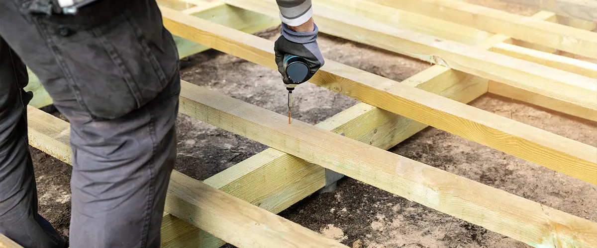 Construction worker using a drill for a wooden deck installation in Farragut, framing on the ground, close-up on hands and tool.