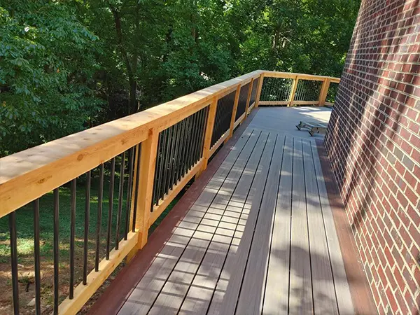 Modern wooden deck with natural wood railing and black balusters, adjacent to a brick wall, overlooking a lush green backyard.