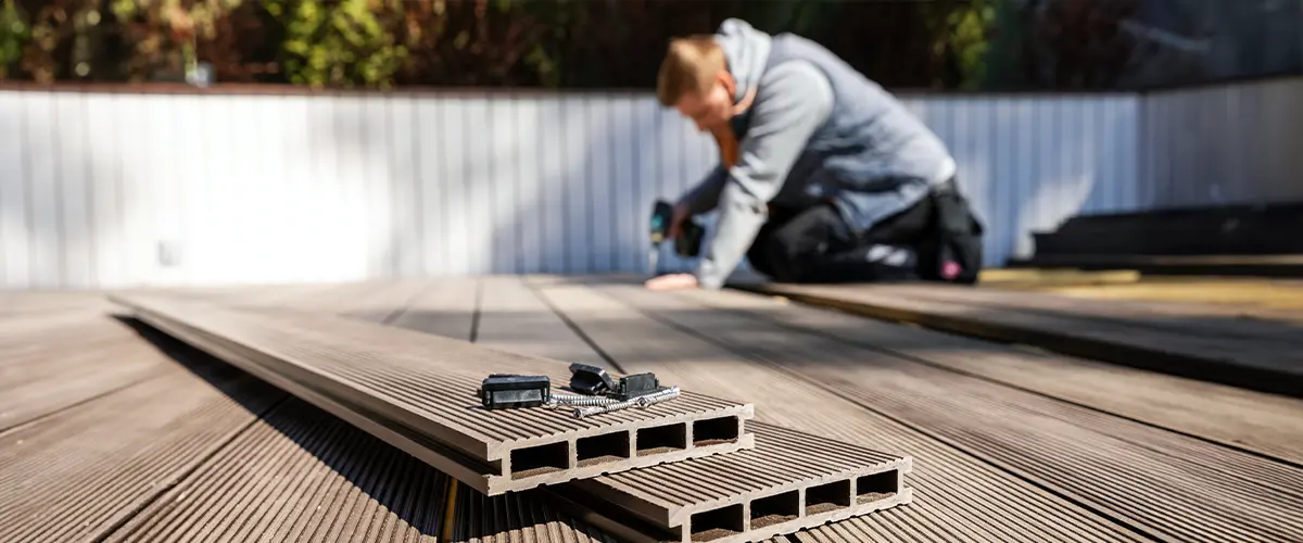 Close-up of composite decking boards with fasteners, screws, and a person repairing a deck in the background using a drill.