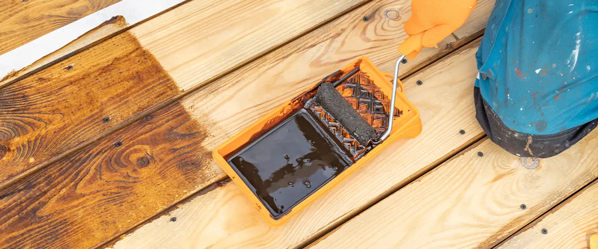 Worker applying wood stain to a deck using a paint roller during a refinishing project - deck staining and restoration service. Solid Deck Stain vs. Semi-Transparent