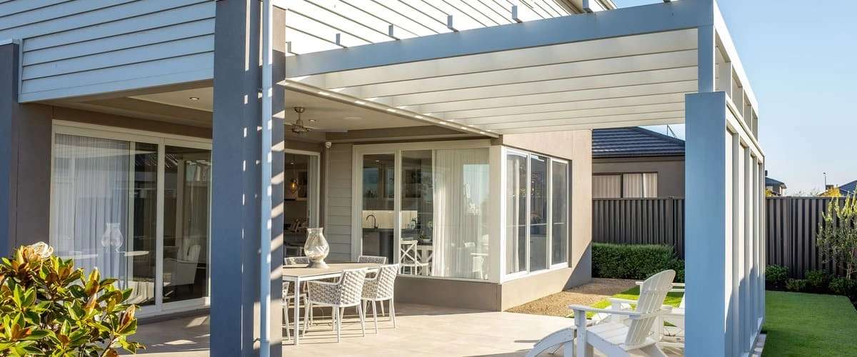 A modern white motorized louvered pergola covering an outdoor dining area with a white table, woven chairs, and Adirondack seating on a sunny backyard patio.