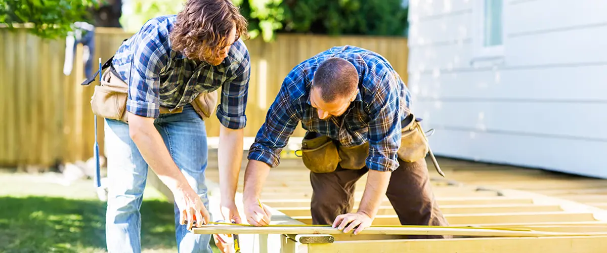 Two professional builders measuring and cutting wood while constructing a deck in a sunny backyard.