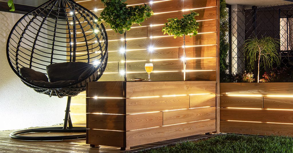Contemporary patio with hanging egg chair, illuminated wood privacy wall, and lush greenery accents.