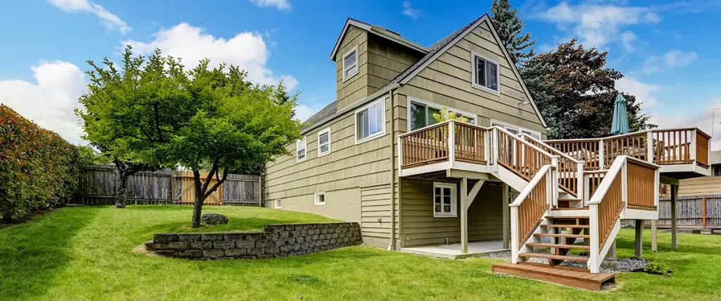 Backyard with elevated wooden deck, staircase, and landscaped lawn around green house under blue sky.