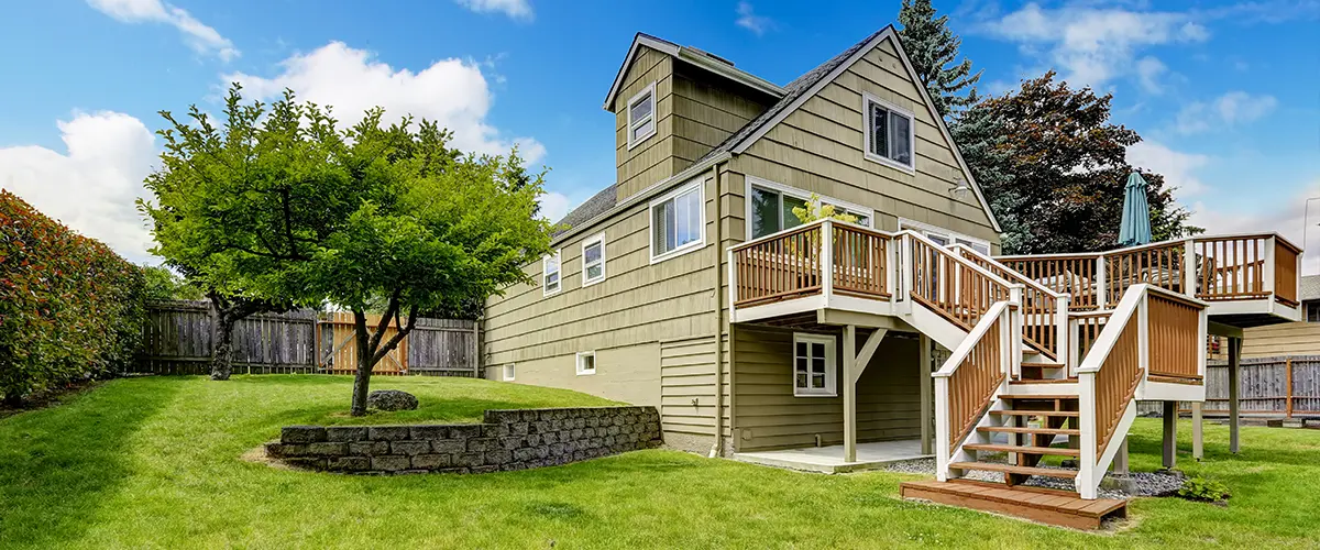 Backyard with elevated wooden deck, staircase, and landscaped lawn around green house under blue sky.
