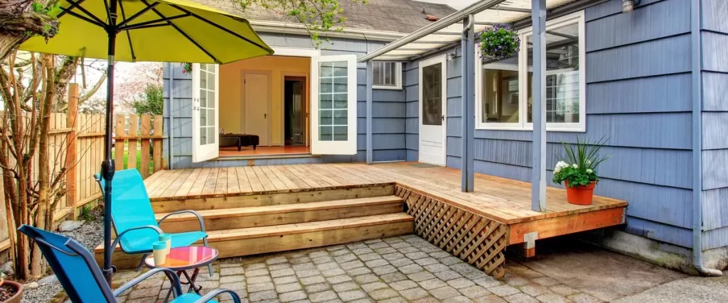 A small elevated wooden deck attached to a blue clapboard house, featuring wide stairs leading down to a stone paver patio with turquoise lounge chairs and a lime green umbrella.