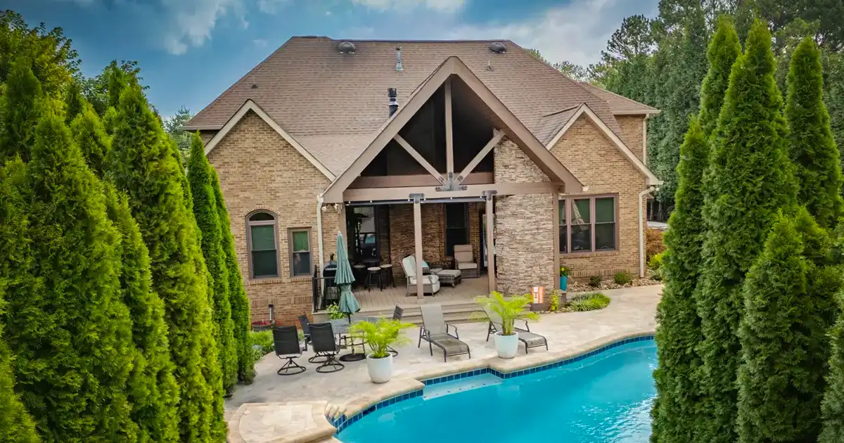 Luxury backyard with inground pool, covered patio, and brick home surrounded by tall privacy trees and outdoor seating - featured