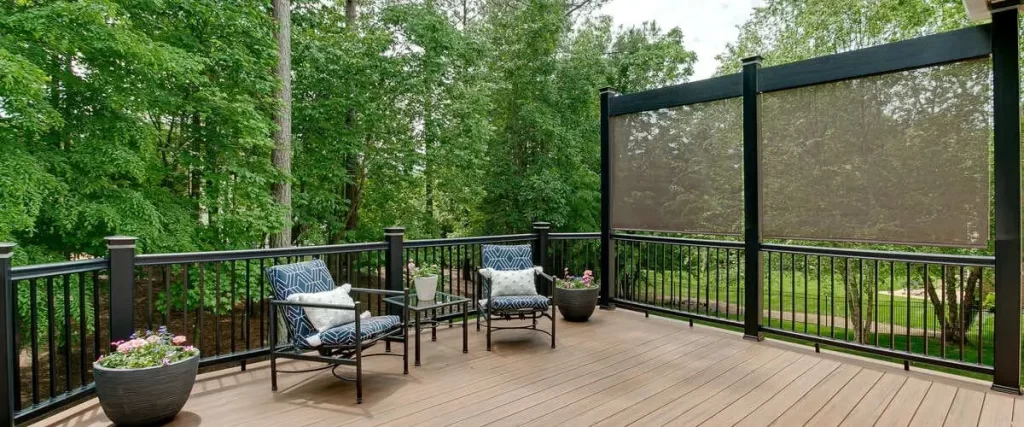 Two blue geometric patterned outdoor chairs with white pillows sit on a brown composite deck featuring black metal railings and a tall brown privacy shade screen overlooking a lush green forest.