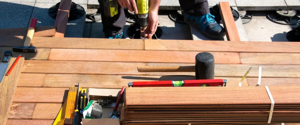 Person installing wooden decking using a power drill, surrounded by tools like a hammer, mallet, spirit level, and a box of hardware