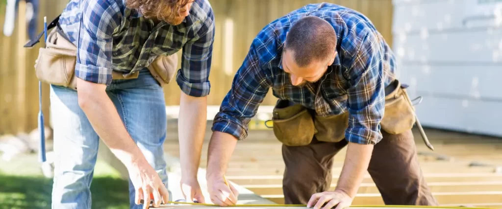 Two workers in plaid shirts and tool belts measuring a wooden plank outdoors