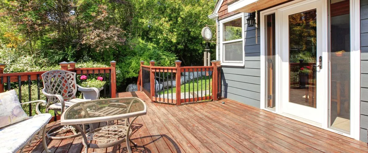 Wooden deck attached to a dark blue house with white trim, featuring a glass-top table, metal chairs, and a cushioned bench