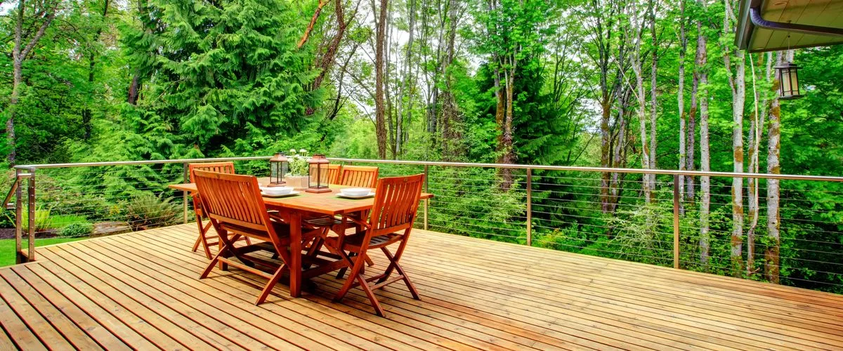 Wooden deck set in a forested environment, featuring a metal railing with horizontal cables and a wooden dining table surrounded by six chairs