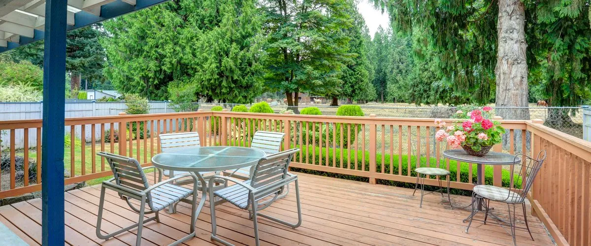 Wooden deck attached to a house, featuring a round glass-top table with four metal chairs and a smaller table with a flower arrangement and ornate chairs