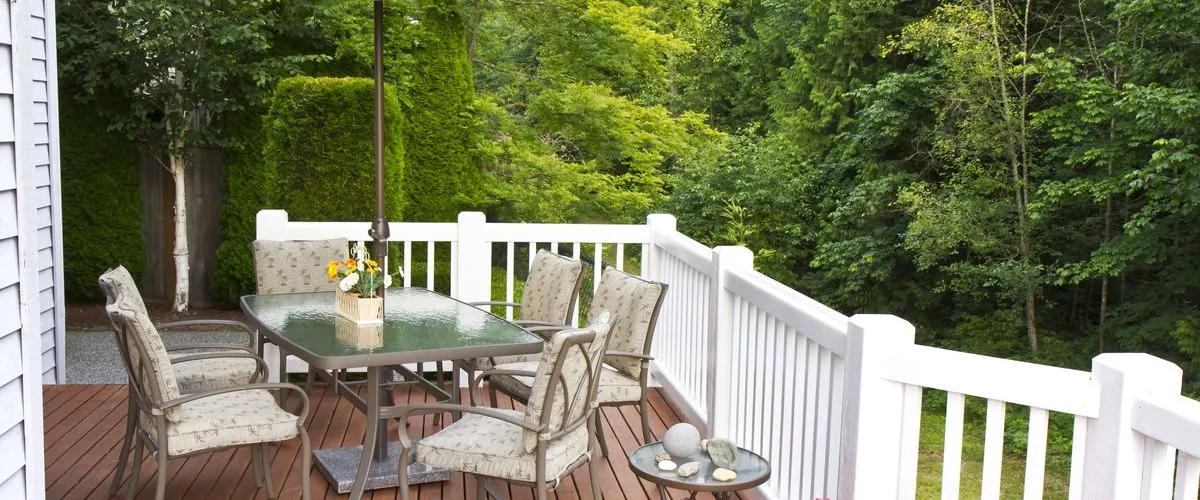 Outdoor deck with white railings overlooking a lush forest, featuring a glass-top dining table with six cushioned chairs and a large umbrella for shade