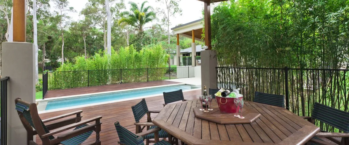 Covered outdoor patio with a wooden deck beside a small rectangular pool, featuring a round wooden table surrounded by cushioned chairs