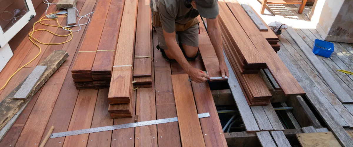Person kneeling on a wooden deck under construction, measuring a plank with a tape measure