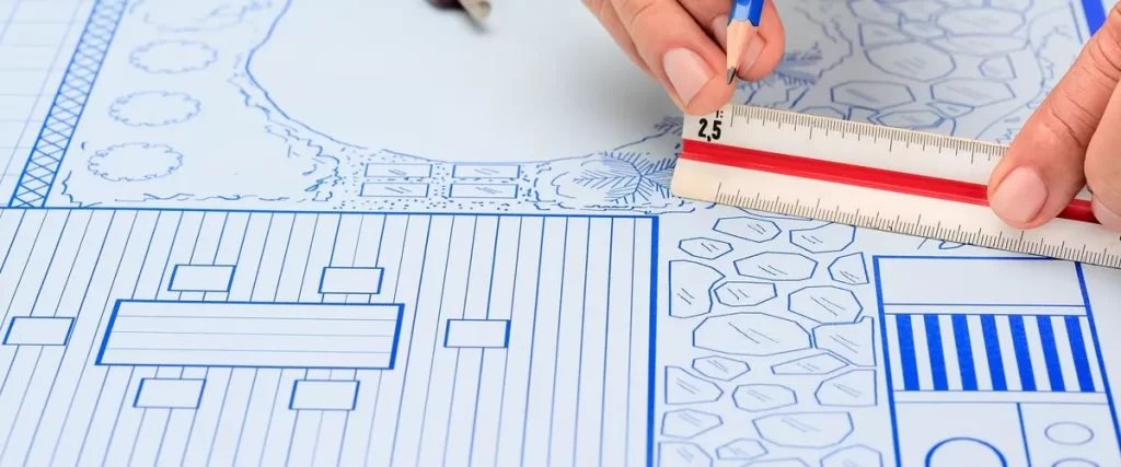 Hands holding a pencil and a ruler, working on a detailed architectural blueprint or plan for an outdoor space, possibly for deck inspection.