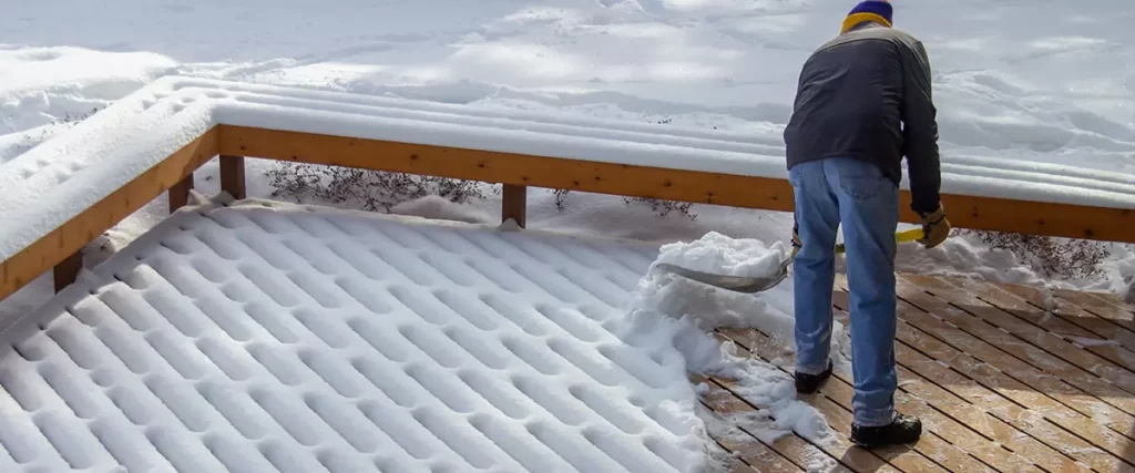 A person in jeans and a jacket is using a shovel to clear a deep layer of snow from a wooden deck with railing during winter.