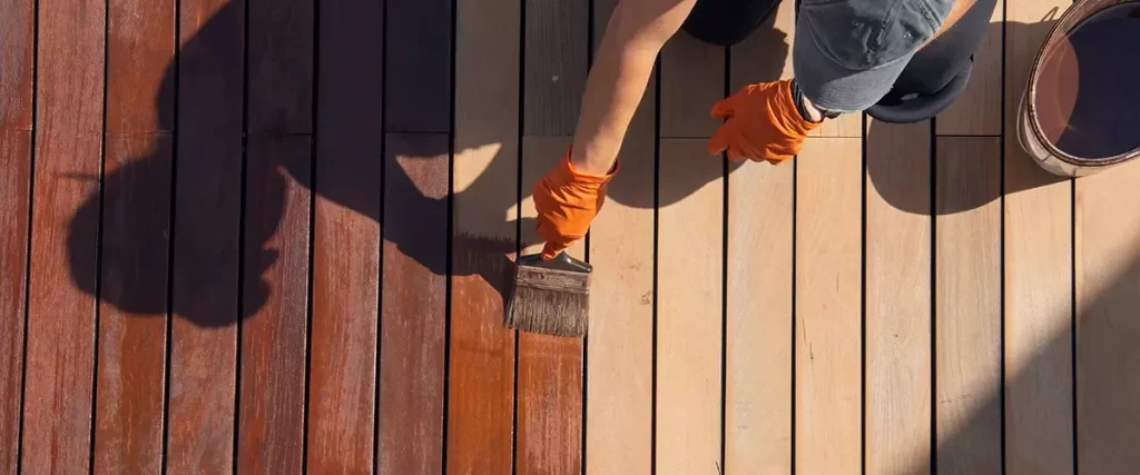 Overhead shot of a person wearing orange gloves applying a reddish-brown stain to light-colored wooden deck planks using a wide paintbrush.