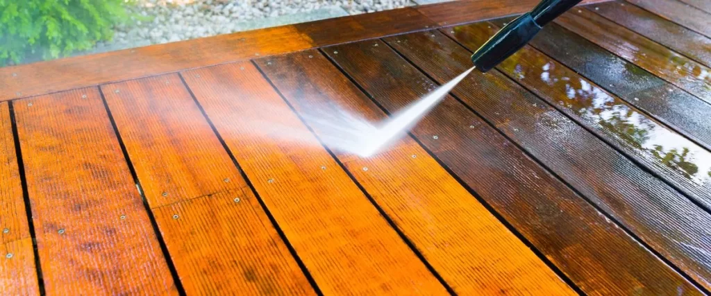 Close-up shot of a dark brown wooden deck being cleaned with a high-pressure water spray, showing the contrast between the clean and dirty wood planks.