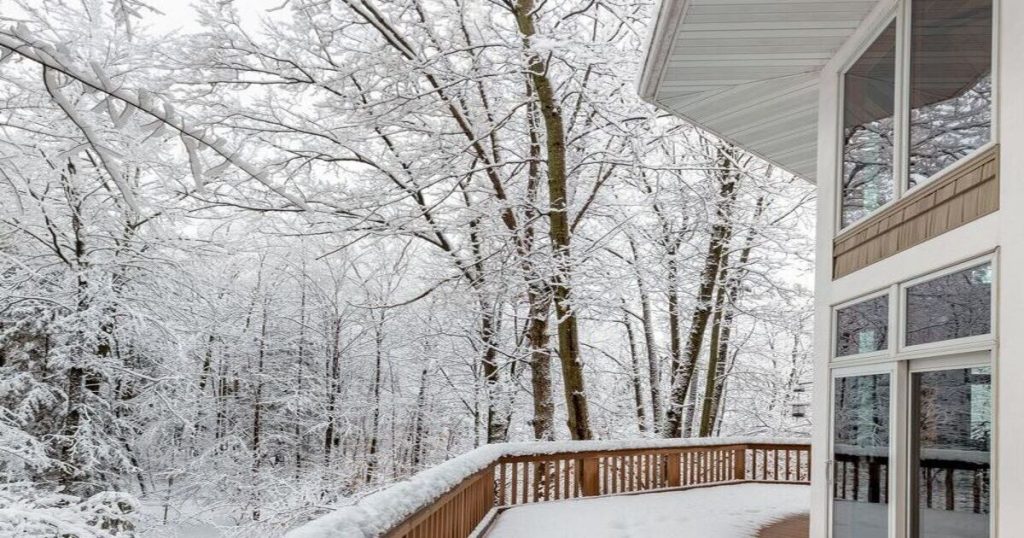 Exterior view of a wooden deck with a railing, completely covered in snow, looking out onto a forest of trees also coated in a fresh layer of snow.