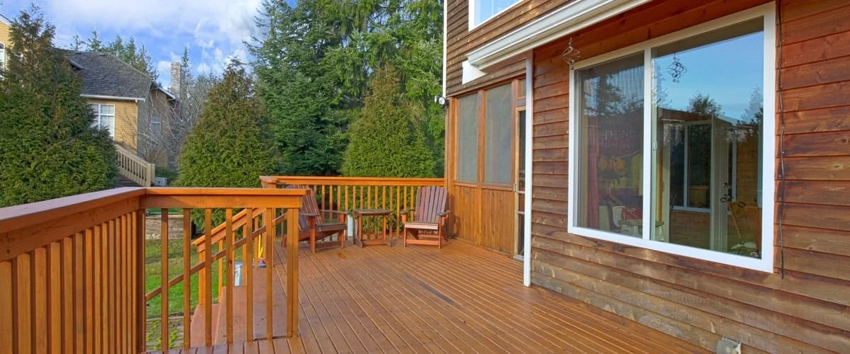 A spacious polished cedar wood outdoor deck featuring traditional wooden railings, a short flight of stairs leading to a lawn, and two matching Adirondack chairs with a small side table against a rustic wood-sided house.