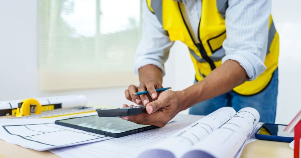 A construction professional in a high-visibility vest using a calculator and digital tablet to estimate costs and requirements for obtaining deck permits based on architectural blueprints.