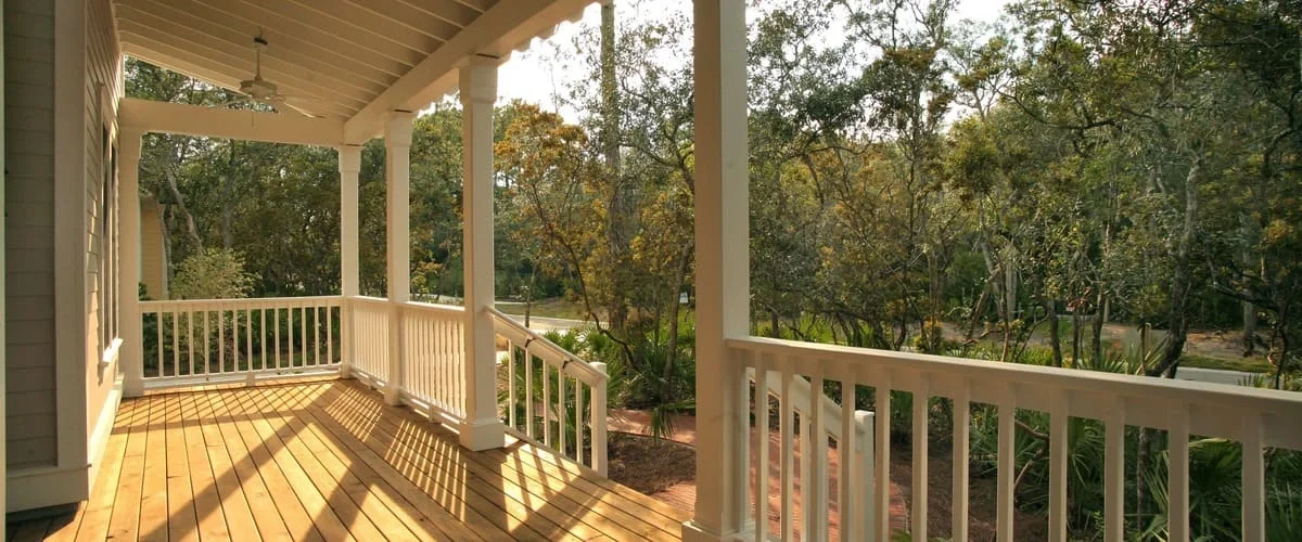 A sunlit covered porch featuring light natural wood floorboards, crisp white railings and support pillars, a ceiling fan, and a view of a lush garden pathway with various trees and tropical plants.