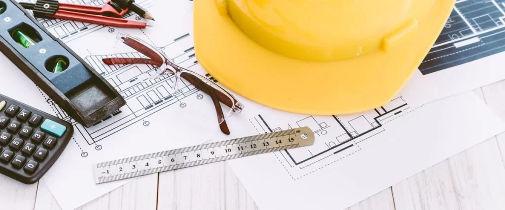 A flat lay of engineering tools including a yellow hard hat, ruler, level, and calculator sitting on top of site plans used to secure deck permits.