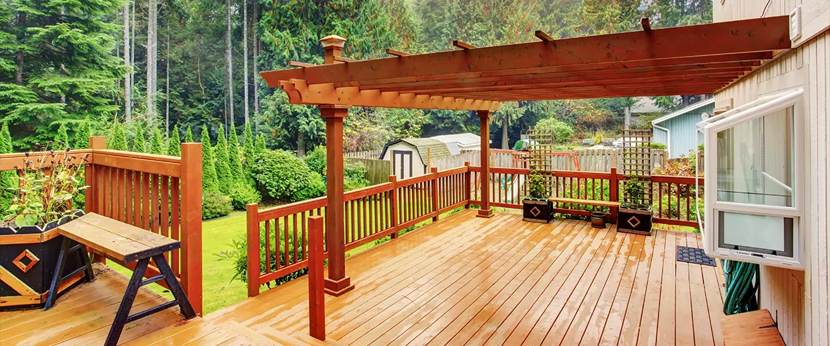 A spacious, wet cedar wood deck featuring a large overhead pergola, matching wooden railings, and black decorative planters overlooking a lush green backyard with evergreen trees.