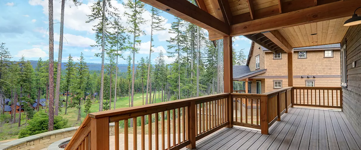 A spacious elevated residential deck with a wood timber frame and railing, offering a panoramic view of a pine forest and a green golf course under a blue sky.