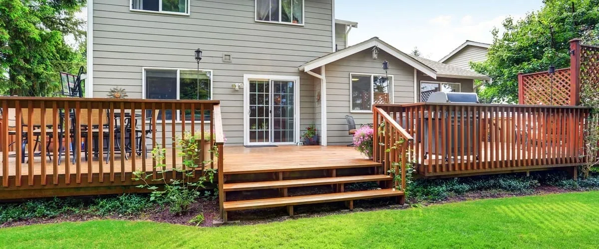 A wide-angle view of a well-maintained multi-level wooden deck featuring redwood stain, vertical slat railings, and wide stairs leading to a green lawn.