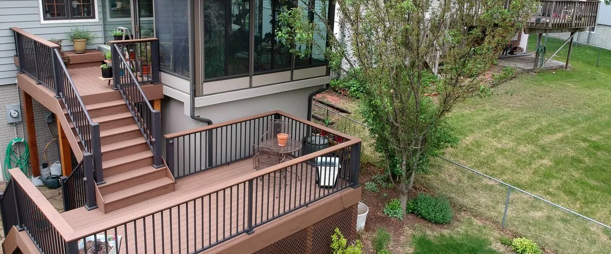 An elevated view of a multi-tiered deck built with brown composite wood and sleek black metal railings, featuring integrated stairs, a small patio table set, and surrounding green lawn and garden landscaping.