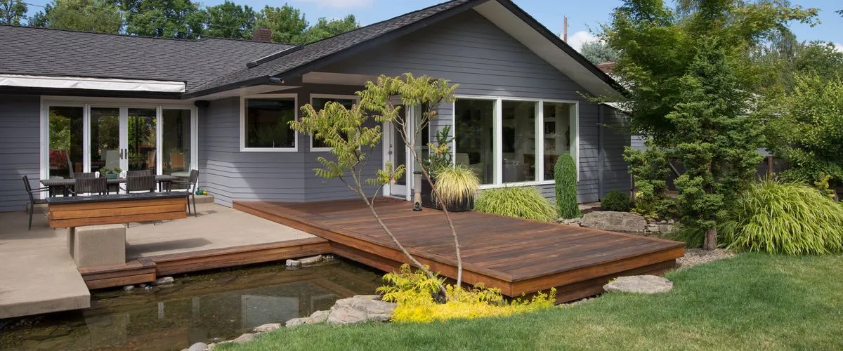 A modern dark-stained wooden floating deck built over a small backyard garden pond, situated next to a grey mid-century modern house with large glass windows and outdoor seating.