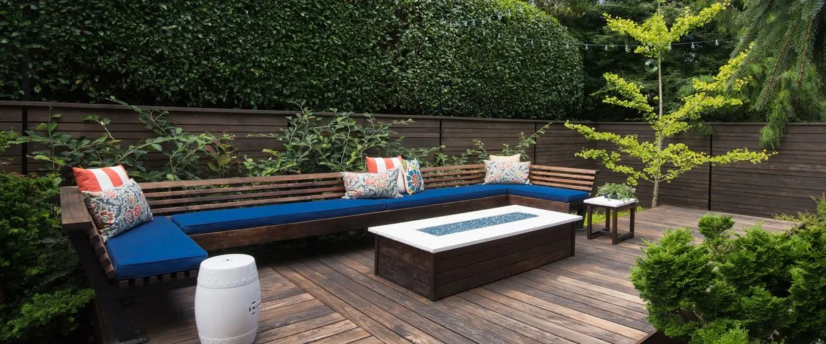 A modern backyard wooden deck featuring a long built-in L-shaped bench with deep blue cushions and colorful accent pillows, centered around a rectangular dark wood fire pit table with a white top, surrounded by a dark horizontal slat fence and lush green privacy hedges.