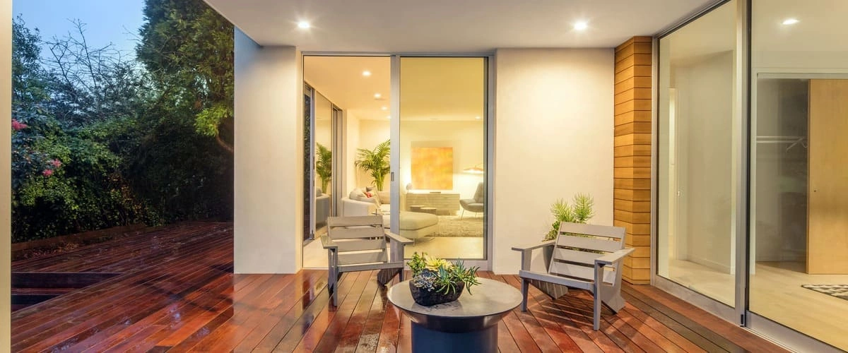 A polished dark wood outdoor deck at dusk featuring two grey contemporary lounge chairs, a circular coffee table with succulents, and a view into a warm, lit modern living room through large sliding glass doors.
