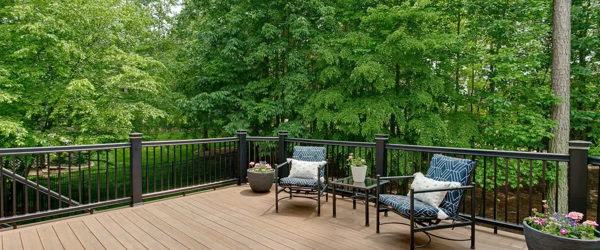 Two blue patterned armchairs with white pillows and a small glass side table on a brown composite deck surrounded by black metal railings and lush green trees.