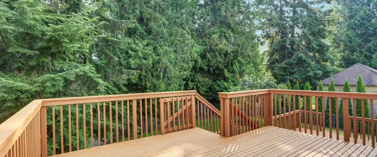 A wide-angle view of a professionally finished redwood wooden deck featuring a secure safety gate, matching railings, and a staircase leading down to a lush green backyard surrounded by dense evergreen trees.