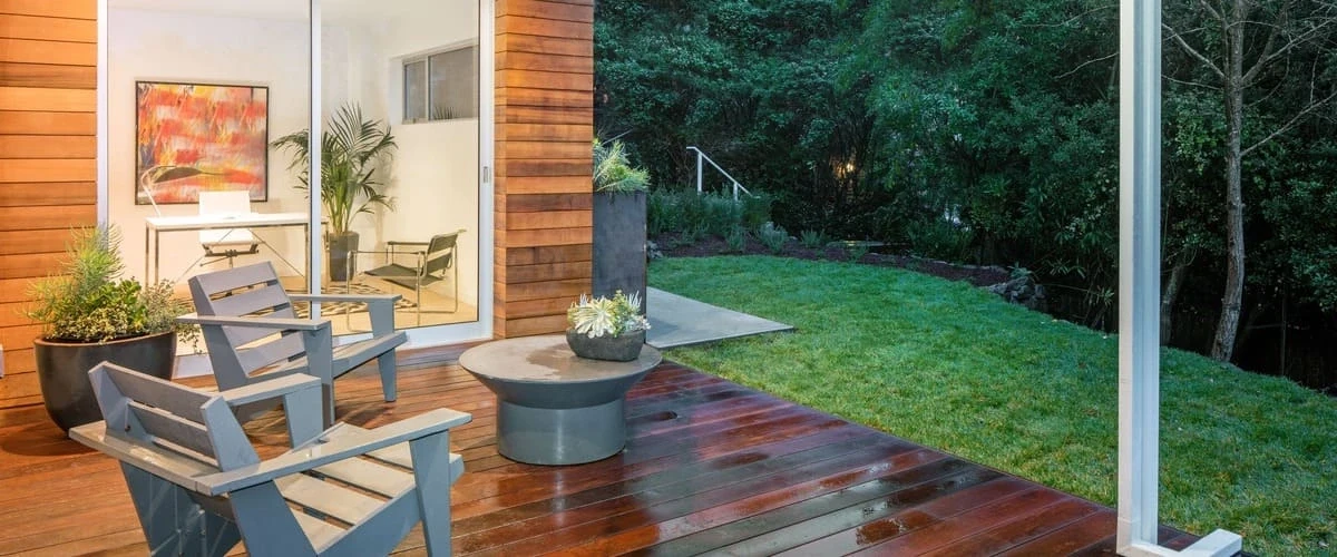 Two modern grey wooden Adirondack chairs and a low circular concrete coffee table arranged on a wet dark wood deck overlooking a lush green lawn and forested backyard at dusk.