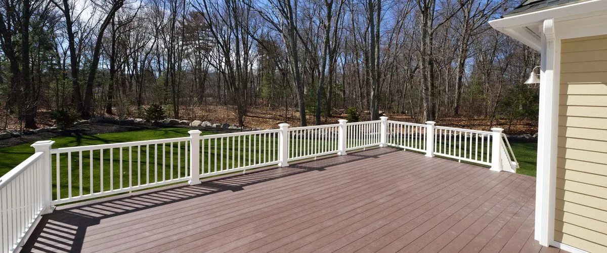 A wide-angle view of a brown composite wood deck featuring white vinyl railings and posts, bordering a lush green lawn and a dense forest background under a clear sky.