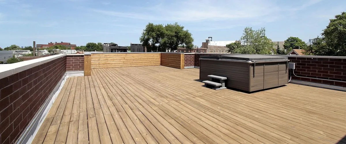 A spacious wooden rooftop deck featuring a large square hot tub with a grey cover and steps, surrounded by a brick perimeter wall under a clear blue sky with a city skyline in the background.