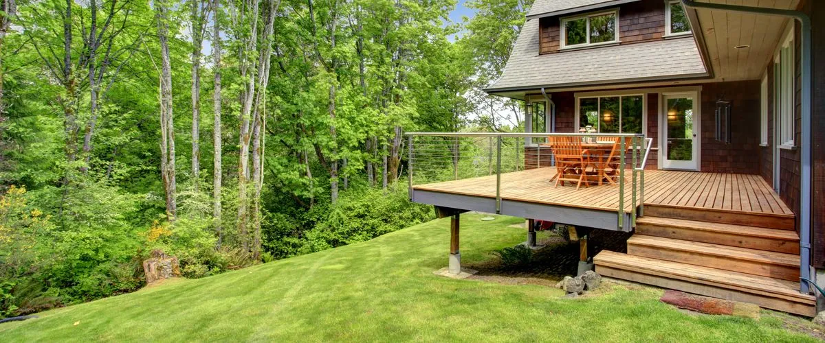 A modern elevated wooden deck featuring sleek stainless steel deck posts and cable railings overlooking a lush green forest with a wooden dining set.
