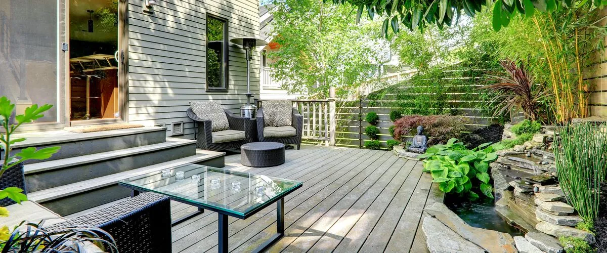 A spacious wooden outdoor deck featuring dark wicker lounge chairs with patterned cushions, a glass-top coffee table, and a tranquil backyard zen garden with a stone waterfall pond, Buddha statue, and lush bamboo fencing.