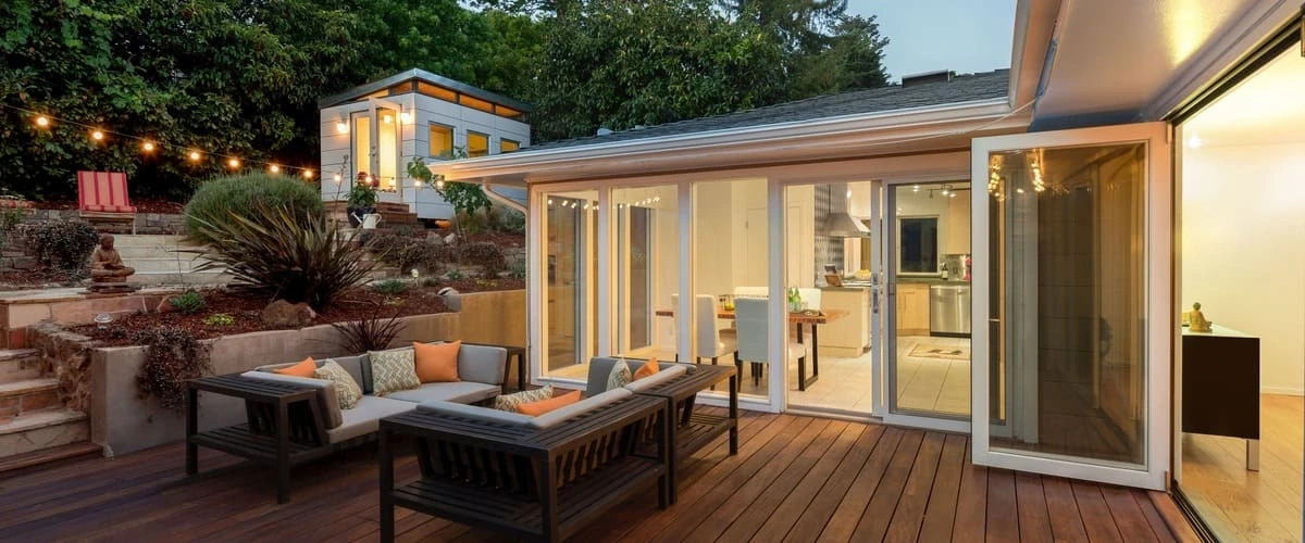 A spacious dark wood outdoor deck featuring a modern grey sectional sofa with orange accent pillows, overlooking a tiered garden with string lights and a small white detached studio building in the background.