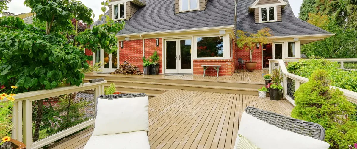 A spacious multi-level tan wooden deck featuring two white wicker lounge chairs in the foreground, overlooking a two-story red brick house with white trim French doors and lush green landscaping.