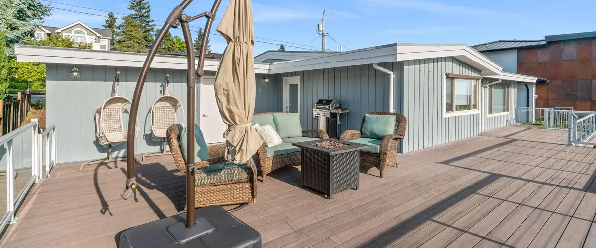 A spacious multi-level wooden outdoor deck featuring a wicker seating area with light green cushions, a square dark brown fire pit table, two hanging bohemian egg chairs against a grey house exterior, and a large tan patio umbrella.