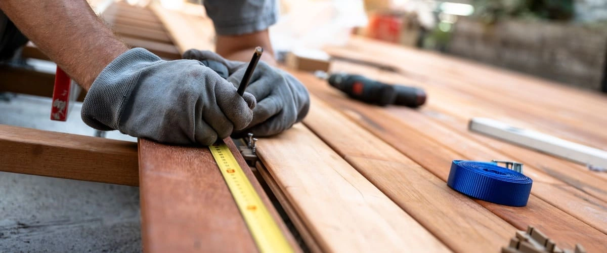 A close-up view of a contractor wearing grey work gloves using a yellow measuring tape and pencil to mark hardwood timber planks during a residential deck installation.